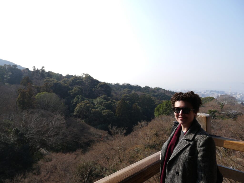 A smiling, non-binary woman in sunglasses stands on a wooden balcony with Kyoto in the distance.